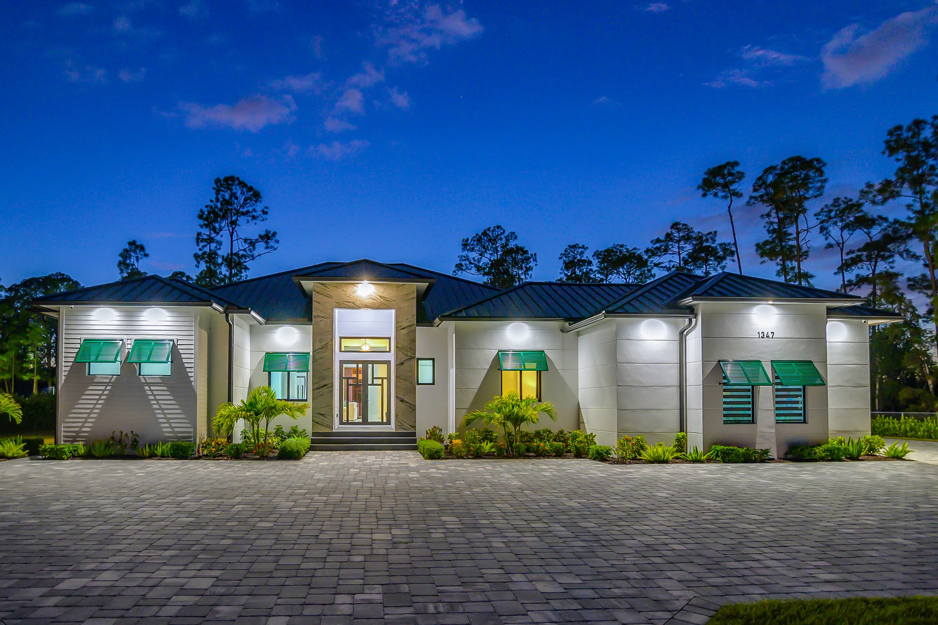 Wide frontal exterior of a modern house at dusk with green window awnings and clear driveway