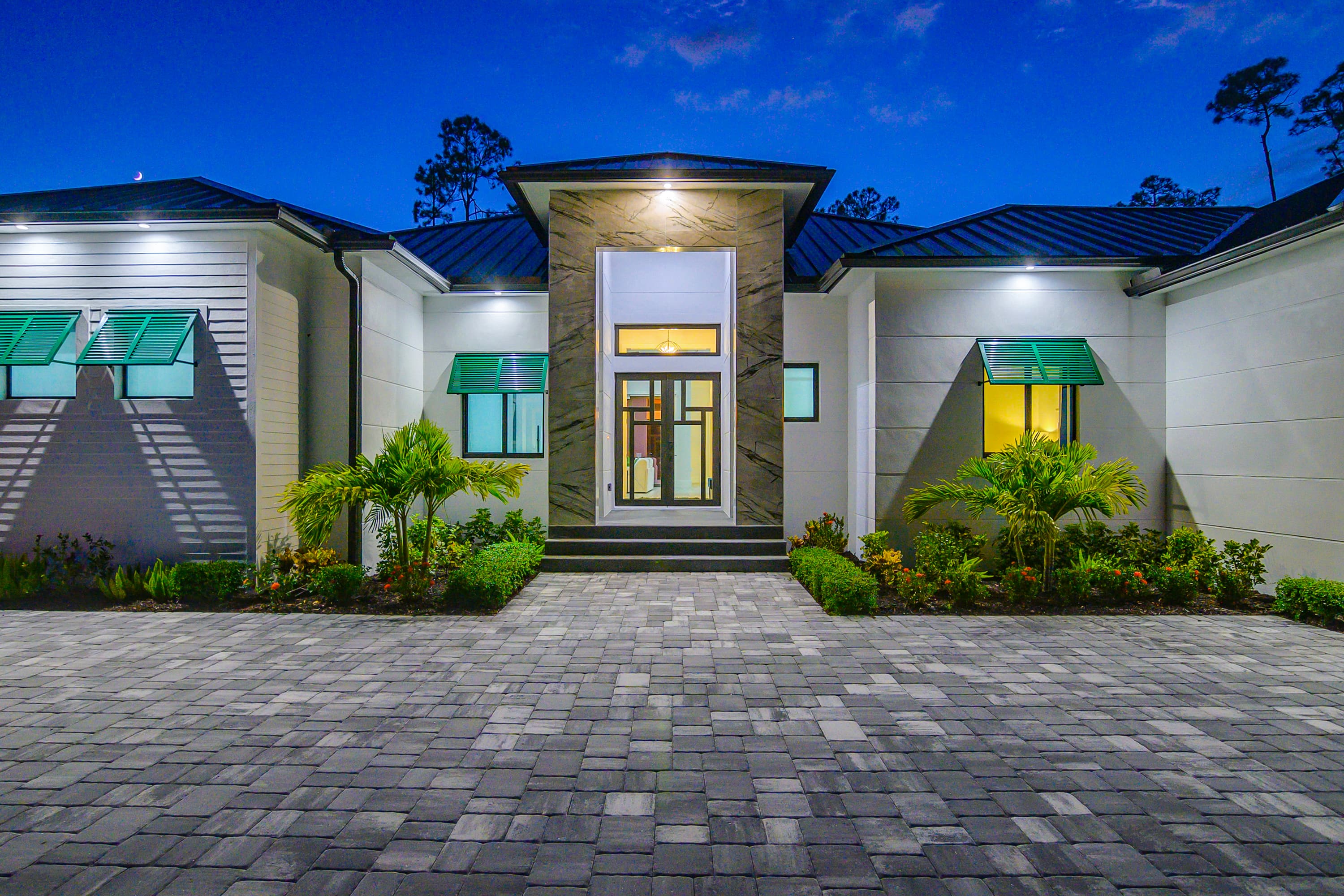 Front entrance of a modern house with glass doors, green window awnings, and lit entrance at dusk