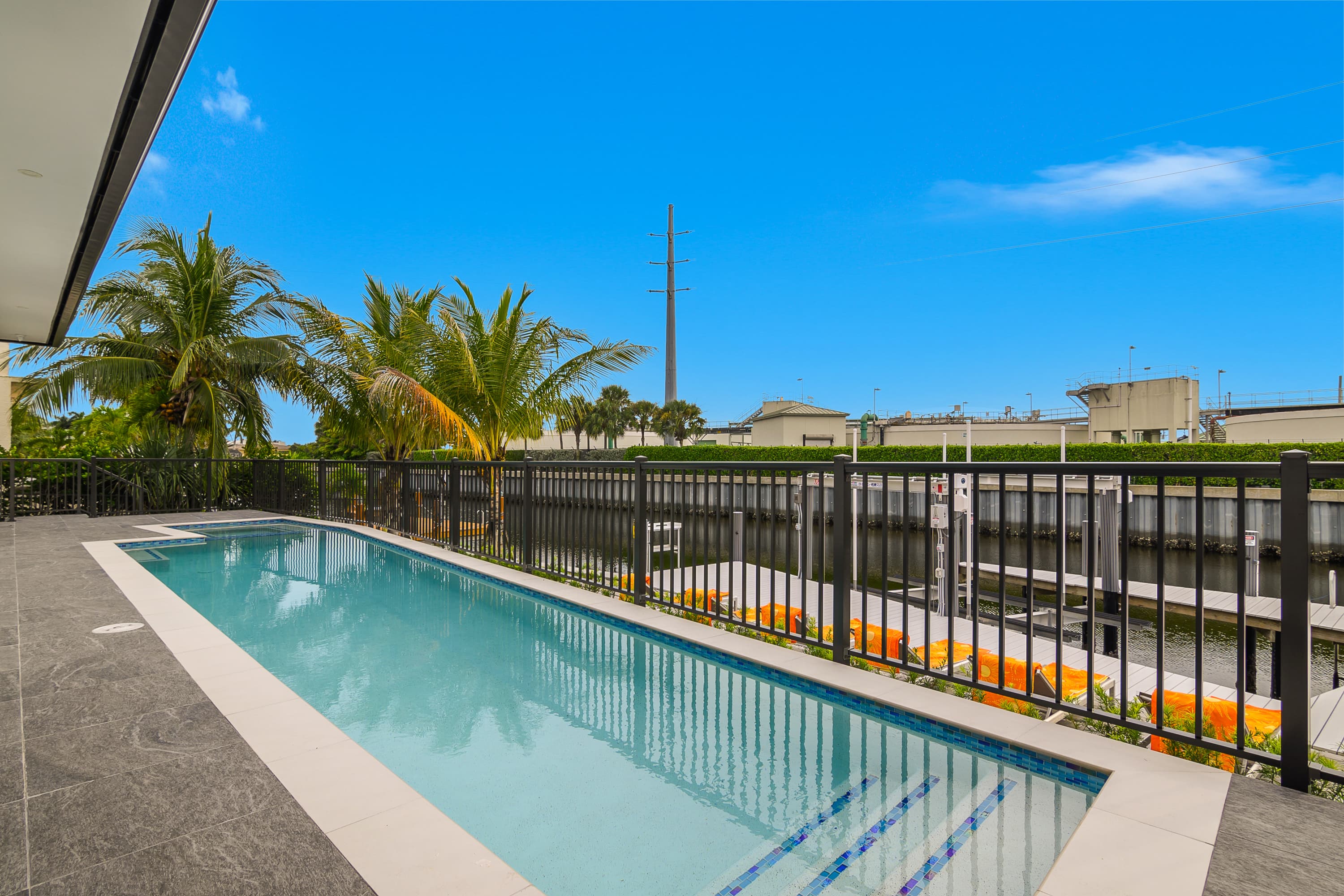 Rectangular pool beside canal with black security fence and palm trees in background under blue sky