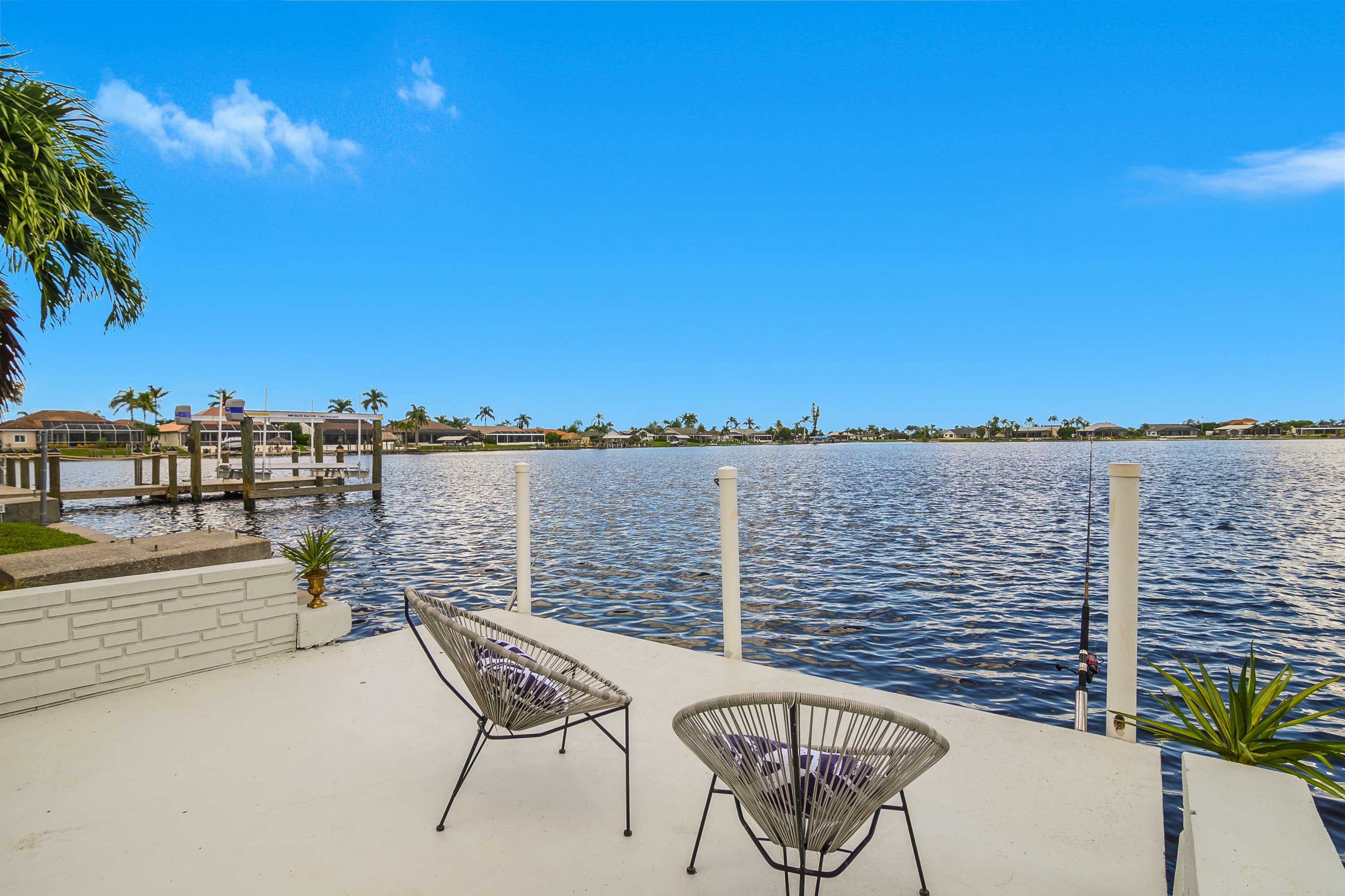 Bright outdoor concrete patio with two chairs and fishing rods facing wide water with houses and palm trees on opposite bank