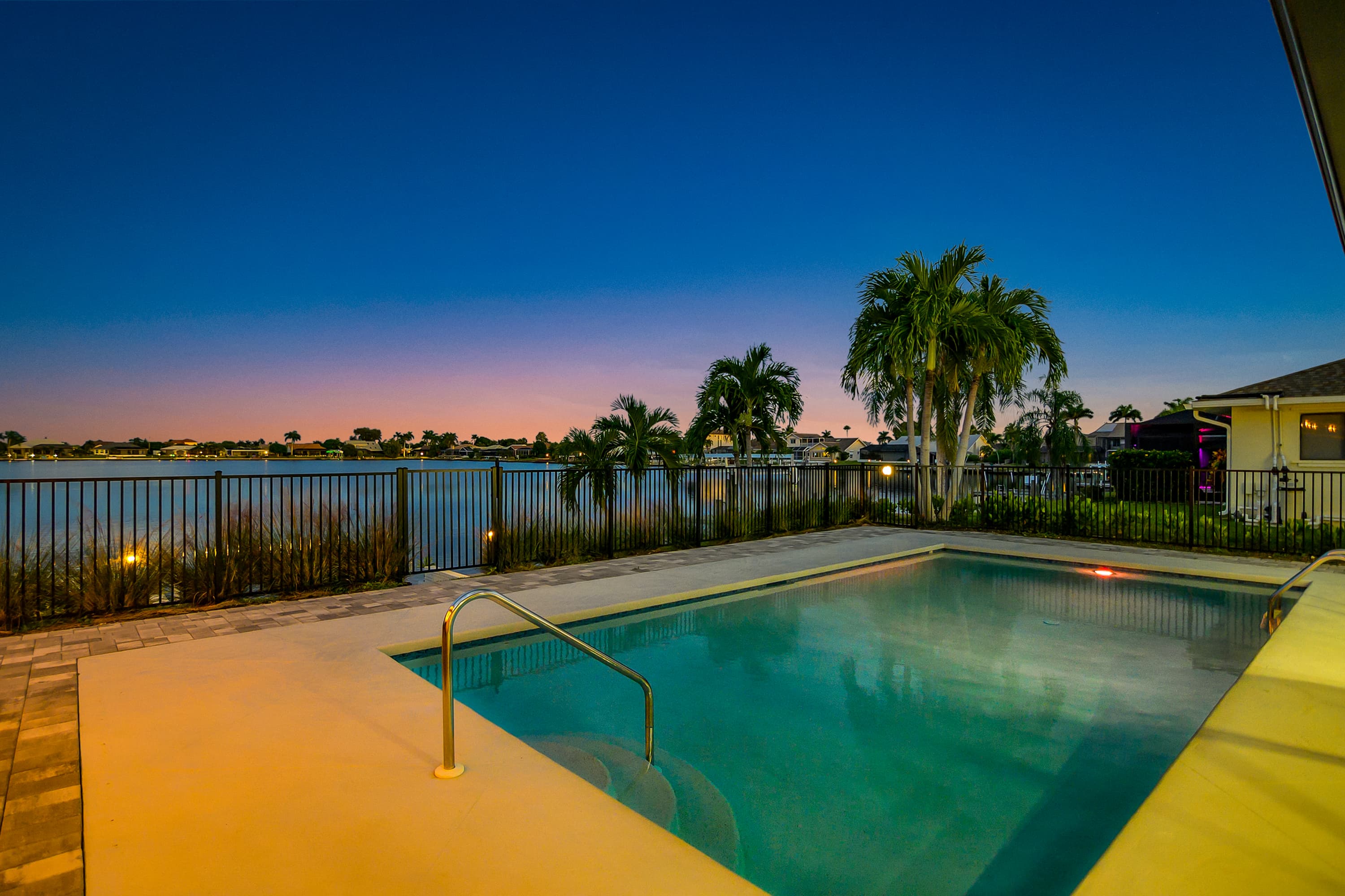 Swimming pool with fence overlooking water and palm trees, lit pergola seating area in background