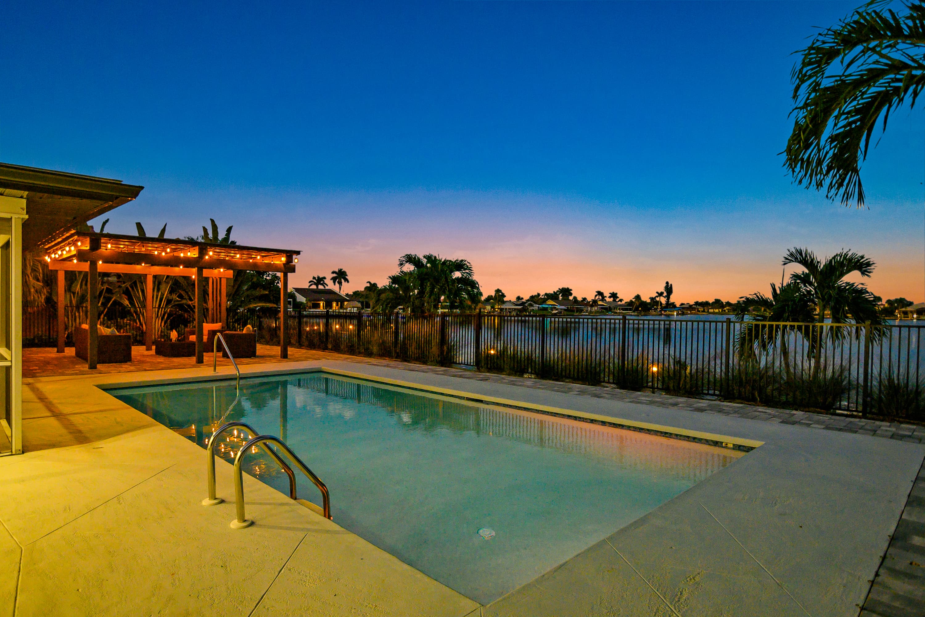 Swimming pool with metal handrails and concrete deck overlooking water and trees at dusk