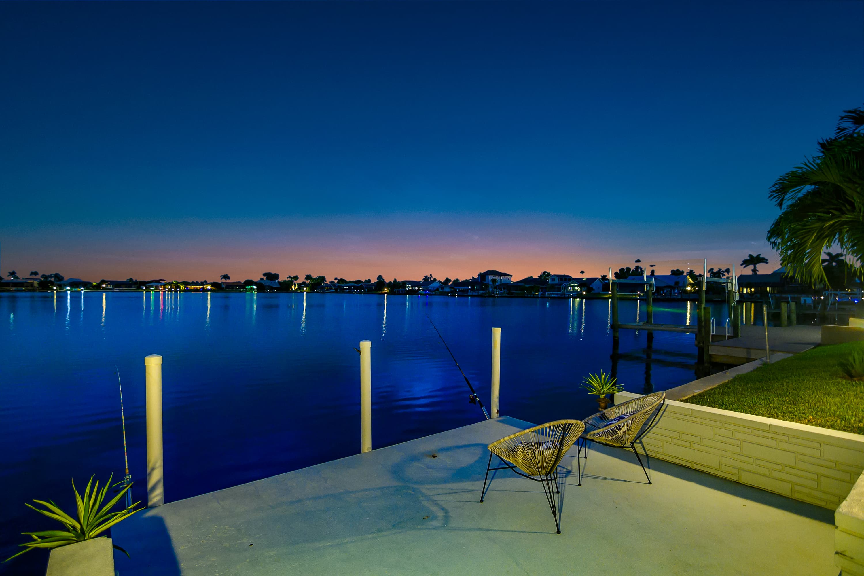Concrete patio with two chairs and fishing rods facing calm water with houses and palm trees across at dawn