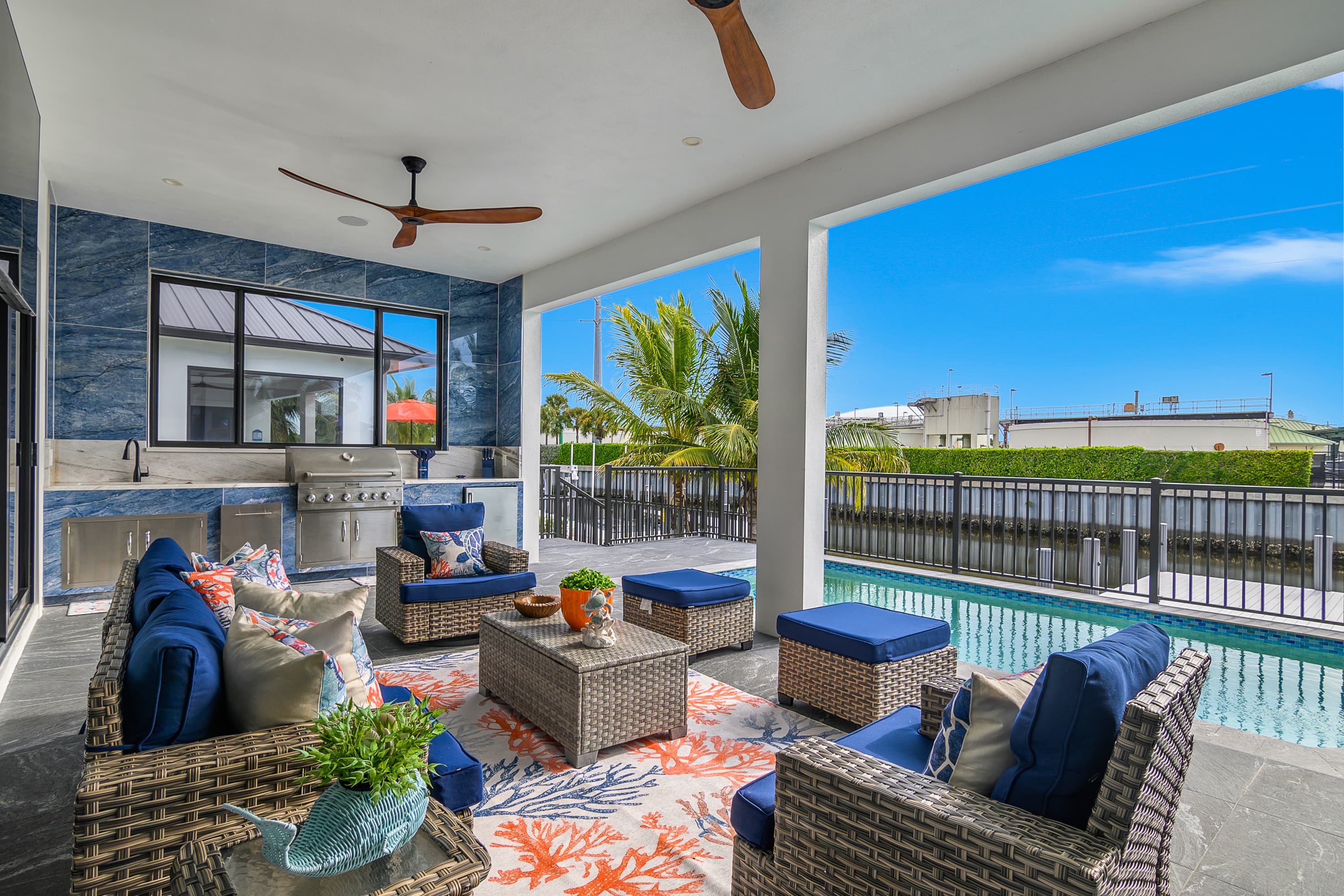 Covered outdoor lounge area with blue cushioned wicker furniture, ceiling fans, a grill, and pool view.