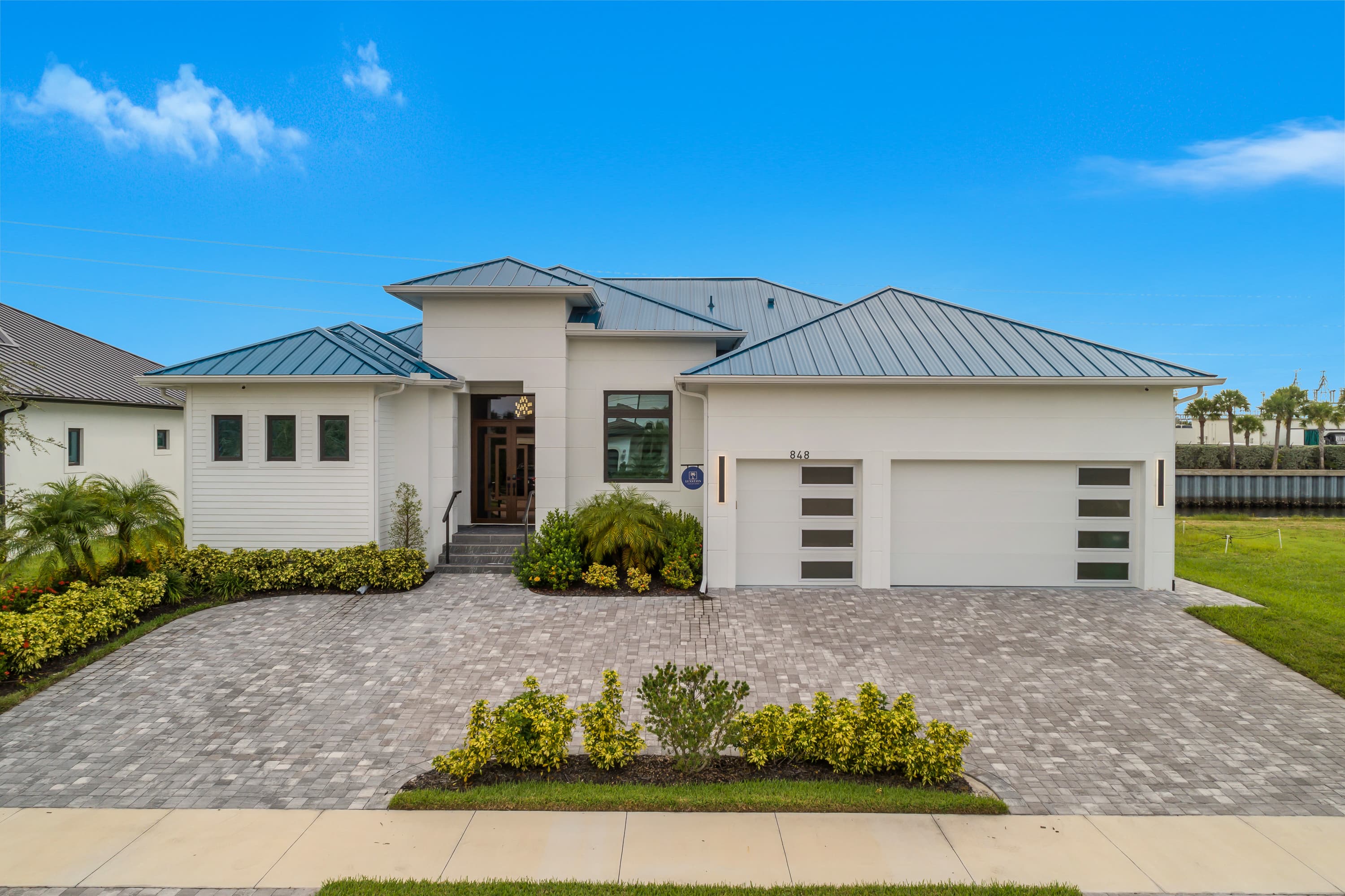 Front exterior of a large modern house with white walls, metal roofs, a driveway, and green landscaping.