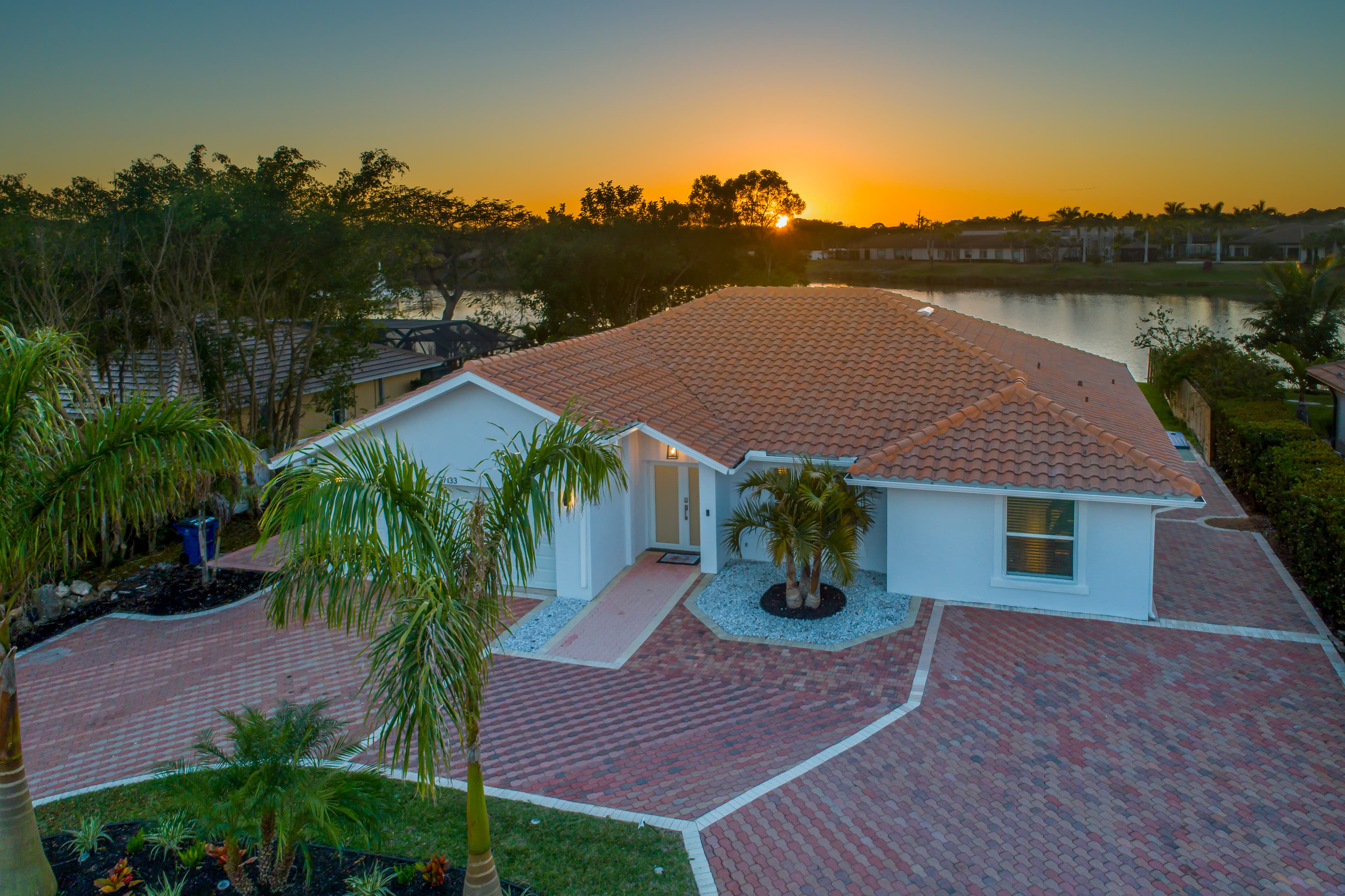 Exterior view of a single-story house with a tile roof at sunset showing driveway, landscaping and lake background