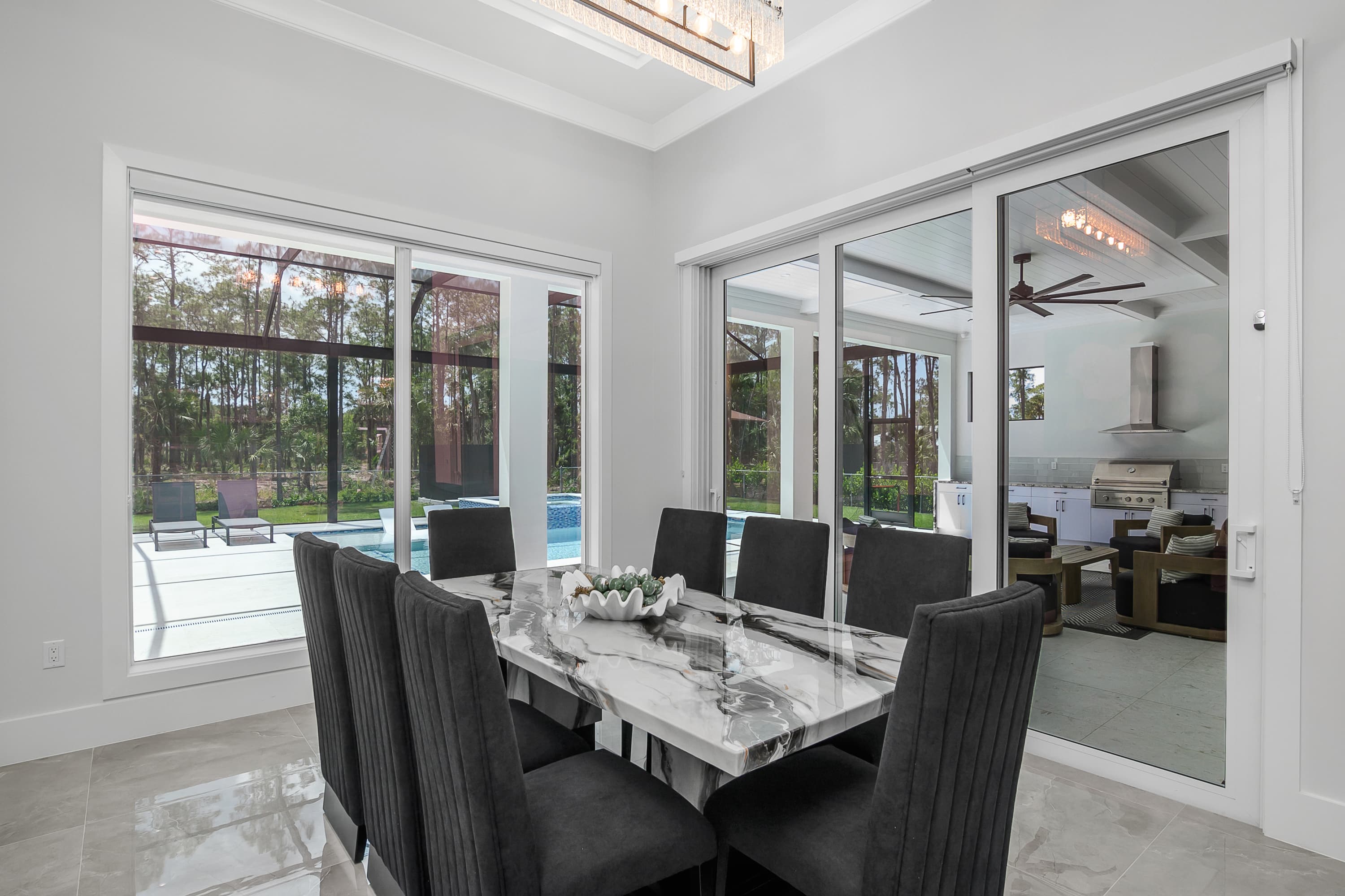 Indoor dining area with marble-top table and black chairs, large windows and sliding doors show pool and outdoor seating